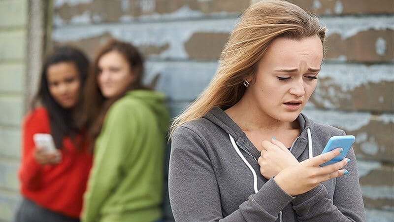 Upset woman experiences cyber bullying over her phone as two snide women look on with phones in hand
