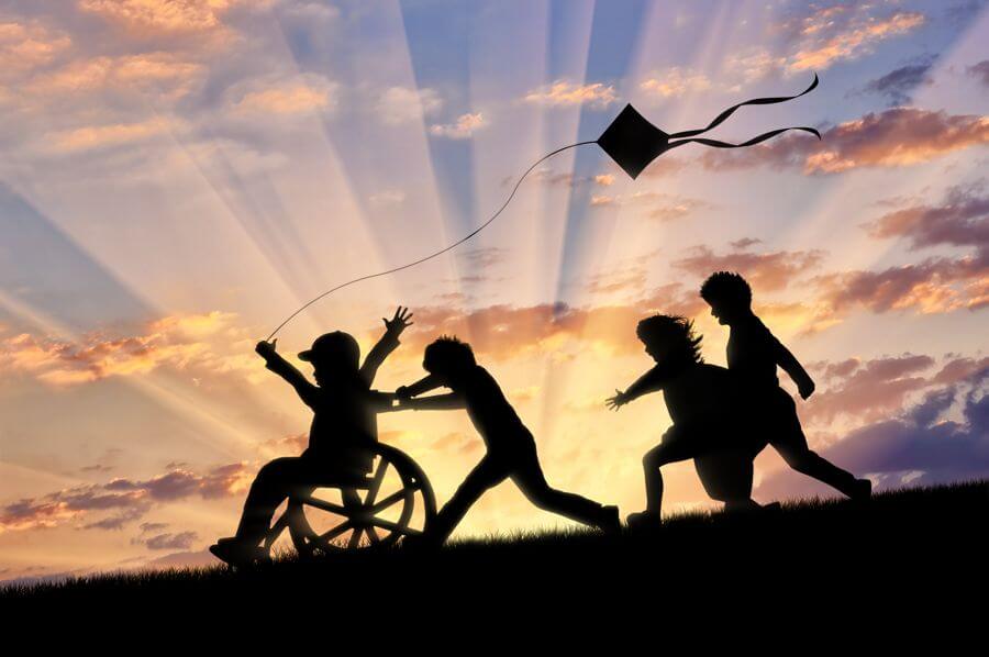 Silhoutted image of playful children pushing a child in a wheelchair and flying a kite